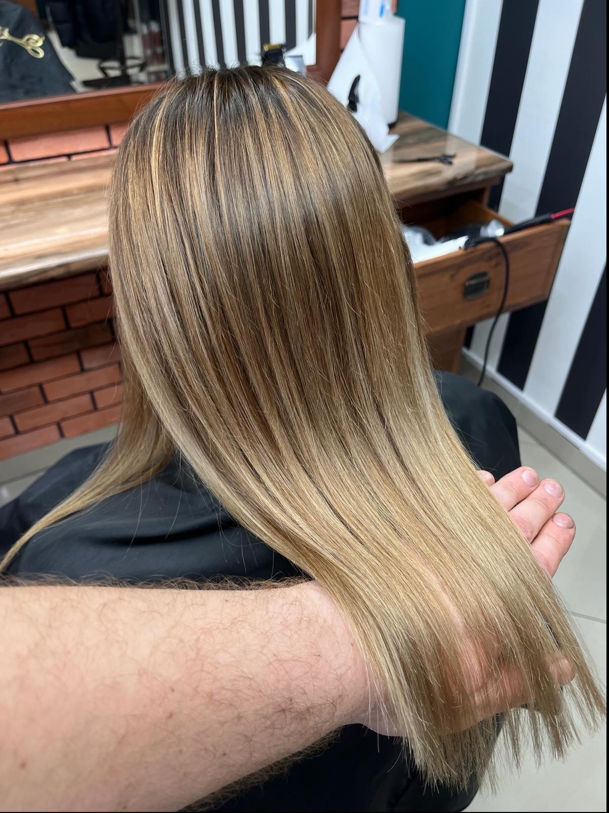 Proste, blond włosy z brązowymi pasemkami, trzymane w dłoni fryzjera, na tle ściany w czarno-białe pasy i ceglanej ściany.