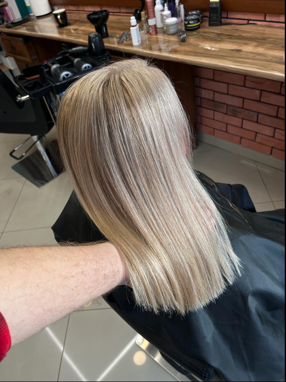 Proste, platynowe blond włosy po koloryzacji, widoczne z tyłu głowy, z ręką stylisty w kadrze i elementami wyposażenia salonu w tle.