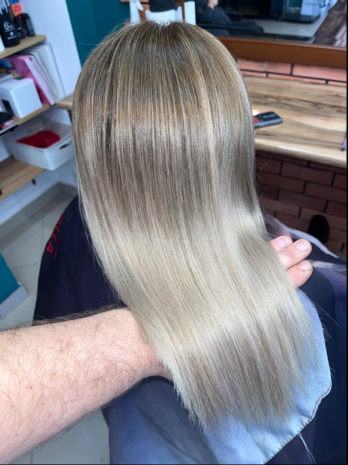 Proste, blond włosy z ombre, widoczne od tyłu, z ręką fryzjera w dolnej części kadru, na tle ściany z cegły w salonie fryzjerskim.