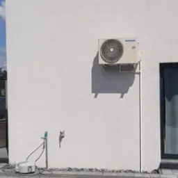 Zewnętrzna jednostka klimatyzacji Panasonic zamontowana na ścianie domu z widocznymi przewodami, obok okno przesuwne, na zewnątrz zadbany trawnik i ogrodzenie.