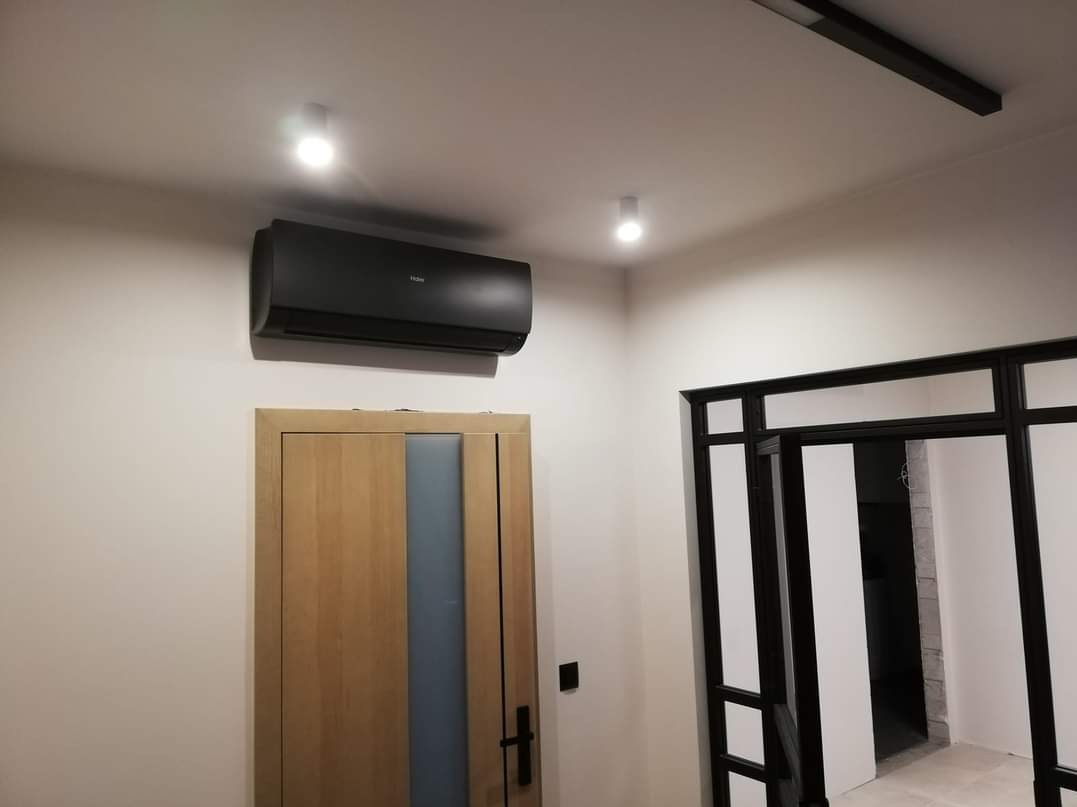 Czarny klimatyzator Haier zamontowany nad drewnianymi drzwiami z pionowym, szarym paskiem szkła, widok z dołu w jasnym wnętrzu z czarną ramą drzwi w tle.
