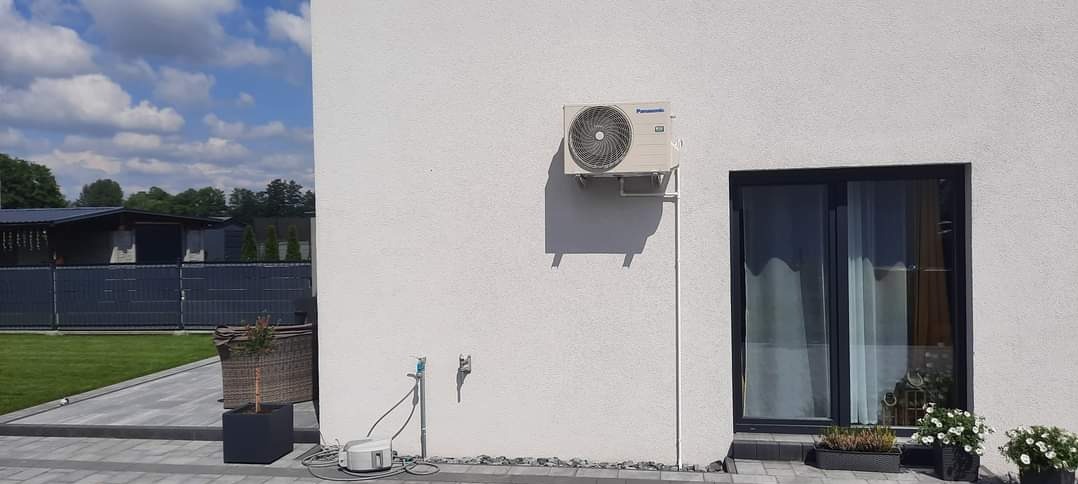 Zewnętrzna jednostka klimatyzacji Panasonic zamontowana na ścianie domu z widocznymi przewodami, obok okno przesuwne, na zewnątrz zadbany trawnik i ogrodzenie.