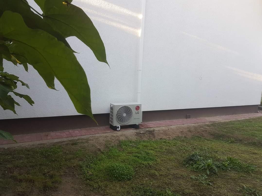 Zewnętrzna jednostka klimatyzacji LG zamontowana przy białej ścianie domu, widok z poziomu trawnika z fragmentem liści drzewa w lewym górnym rogu.