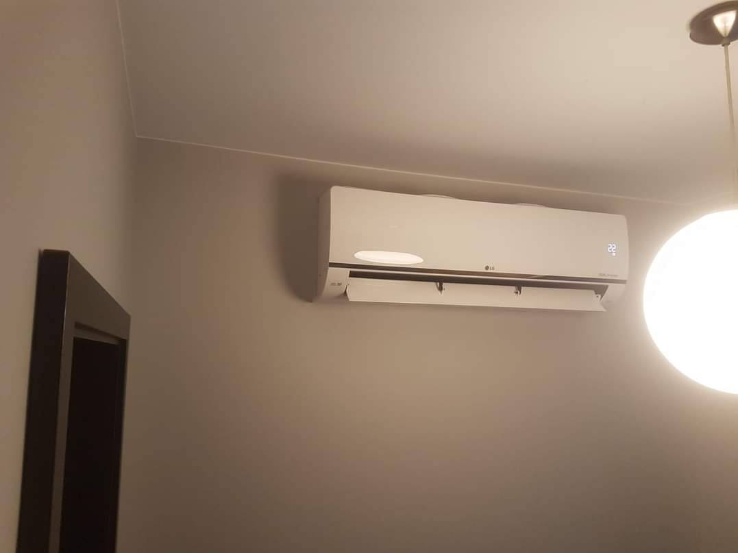 Biały klimatyzator LG zamontowany na szarej ścianie w pokoju, widoczny fragment ramy drzwi i lampa