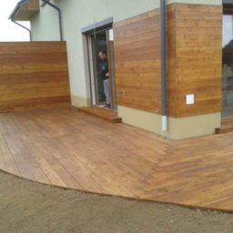 Usługi budowlane - Nowoczesny taras z drewna egzotycznego, z desek ułożonych w jodełkę, przylegający do budynku z drewnianą elewacją i osłoną, widoczny fragment ogrodu.