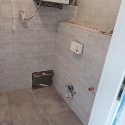 Usługi budowlane - Wnętrze łazienki w trakcie remontu, widoczne płytki imitujące drewno, instalacja grzewcza, niedokończone przyłącza wodno-kanalizacyjne i otwory w ścianach.