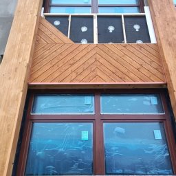 Usługi budowlane - Fragment elewacji budynku z drewnianą konstrukcją i oknami zabezpieczonymi folią, widoczne ocieplenie i montaż desek w jodełkę.