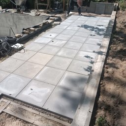 Usługi budowlane - Układanie szarej kostki betonowej na przygotowanym podłożu, widoczny pracownik w czerwonej czapce oraz betoniarka i taczka w tle.