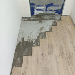 Usługi budowlane - Układanie podłogi z płytek imitujących drewno, widoczny fragment z nałożonym klejem i częściowo ułożone panele, zabezpieczone folią drzwi.