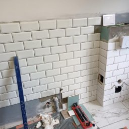 Usługi budowlane - Układanie białych, prostokątnych płytek ściennych typu metro w kuchni, widoczne narzędzia glazurnicze, fragment instalacji wodnej i kanalizacyjnej, oraz zabudowa geberitu w trakcie prac...
