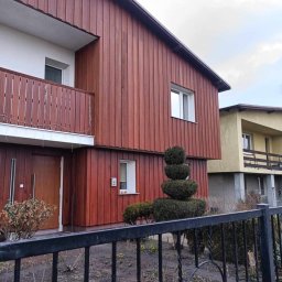 Usługi budowlane - Dwupiętrowy dom z elewacją w odcieniach brązu, drewnianymi panelami pionowymi, balkonem z drewnianą balustradą i ozdobnym krzewem przed wejściem, widziany zza metalowego ogrodzenia.