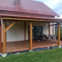 Usługi budowlane - Zadaszony taras z drewna z kompletem mebli ogrodowych. Widoczne elementy konstrukcyjne i wykończeniowe, brązowa podłoga tarasu i fragment domu z czerwonym dachem.