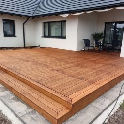 Usługi budowlane - Nowoczesny taras drewniany z dwustopniowymi schodami, wykończony jasnym drewnem, przylegający do białego domu z ciemną stolarką okienną i zadaszeniem.