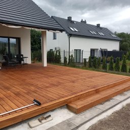 Usługi budowlane - Świeżo wykończony taras z drewna egzotycznego, z widoczną szczotką do zamiatania i młotkiem na obrzeżu, przy nowoczesnym domu z szarą dachówką.