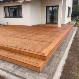 Usługi budowlane - Nowoczesny taras z desek w ciepłym odcieniu brązu, z niskimi schodkami i szarą kostką obrzeżną, na tle białego domu z ciemnymi oknami i zieleni ogrodu.