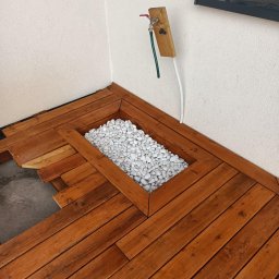 Usługi budowlane - Brązowy taras drewniany w trakcie montażu, z wbudowaną donicą wypełnioną białymi kamieniami, widoczny kran z wężem na ścianie.