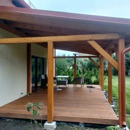 Usługi budowlane - Zadaszony taras z drewnianą konstrukcją i meblami ogrodowymi. Widoczny system rynnowy w kolorze brązowym i kamera monitoringu na ścianie domu.