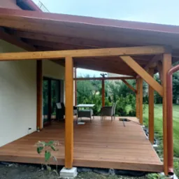 Zadaszony taras z drewnianą konstrukcją i meblami ogrodowymi. Widoczny system rynnowy w kolorze brązowym i kamera monitoringu na ścianie domu.