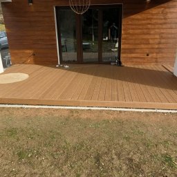 Usługi budowlane - Nowoczesny taras kompozytowy z brązowych desek, przylegający do budynku z drewnianą elewacją. Widoczny fragment trawnika i okrągły dywanik przy drzwiach.