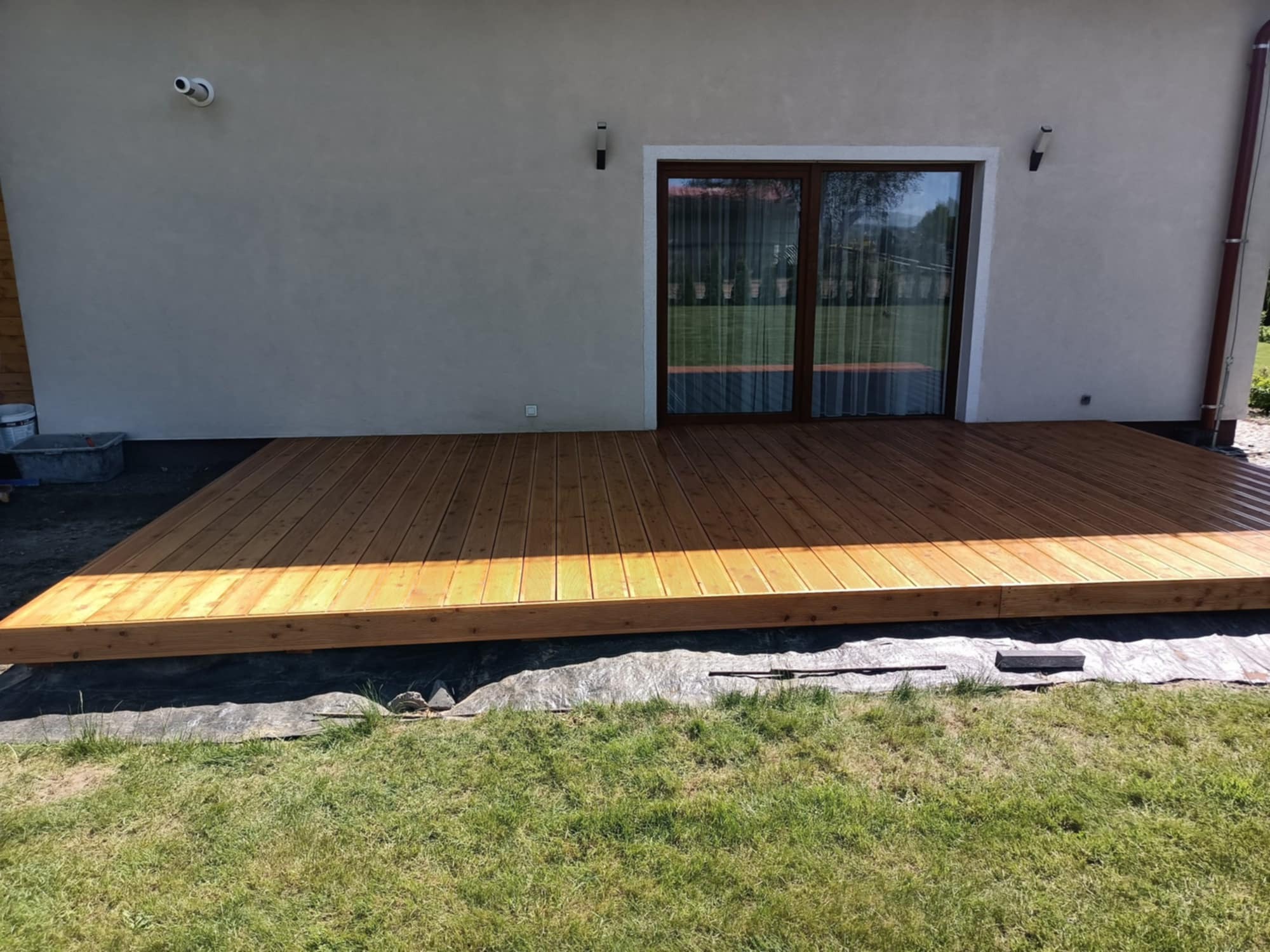 Jasny, drewniany taras przydomowy z widocznymi deskami. W tle ściana budynku z oknem balkonowym. U dołu trawa. Widoczne elementy montażowe.