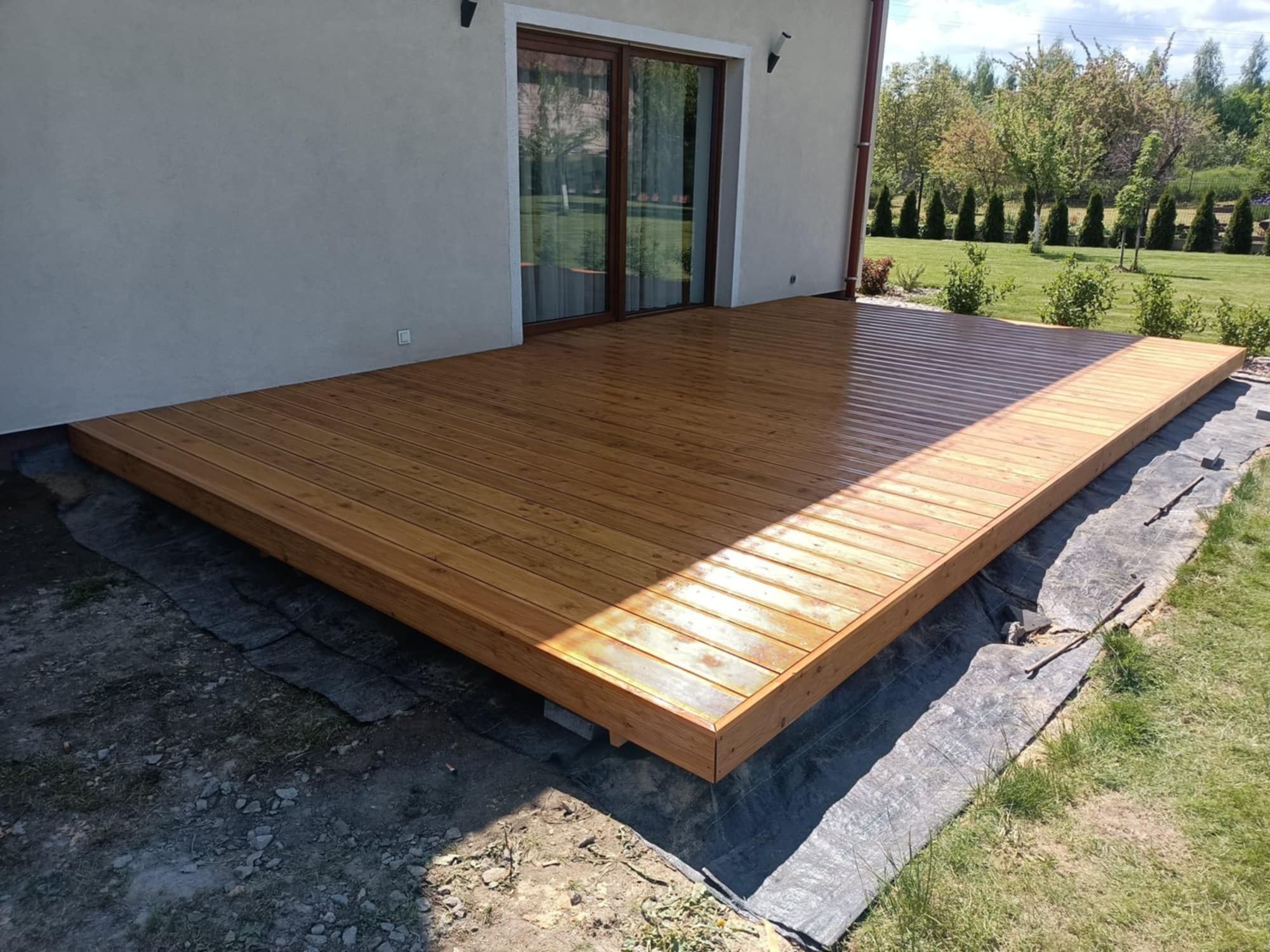 Jasny, drewniany taras przylegający do domu z dużymi, przesuwnymi drzwiami. Widoczne elementy konstrukcyjne i otoczenie z zielenią.