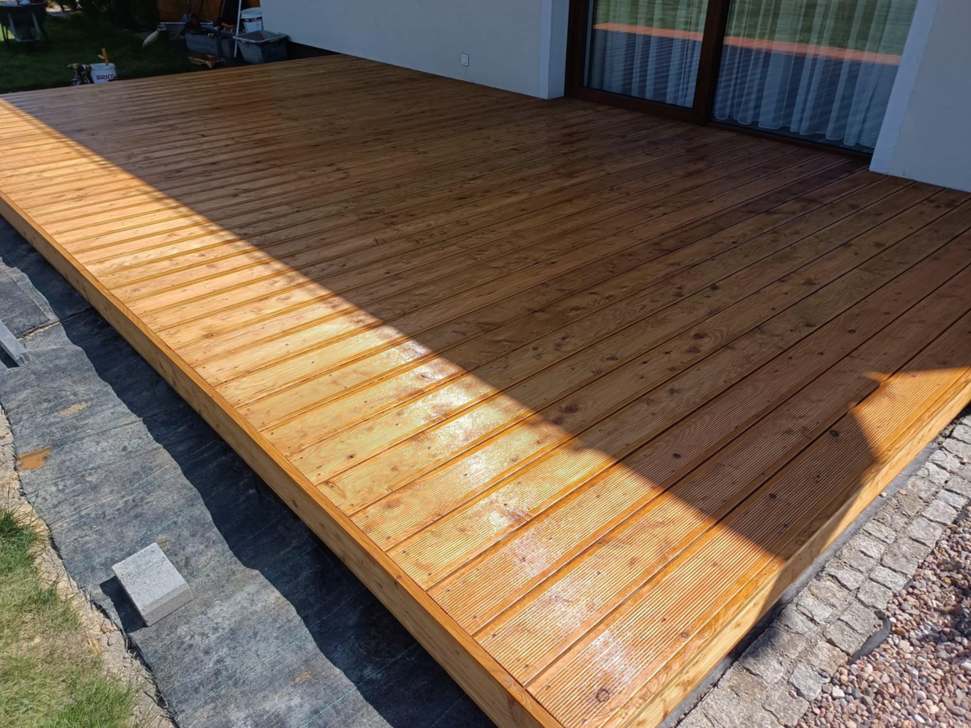 Zaimpregnowany taras z desek drewnianych ułożonych pod kątem, z widoczną strukturą drewna i elementami wykończenia z kostki granitowej. Częściowo zacieniony.