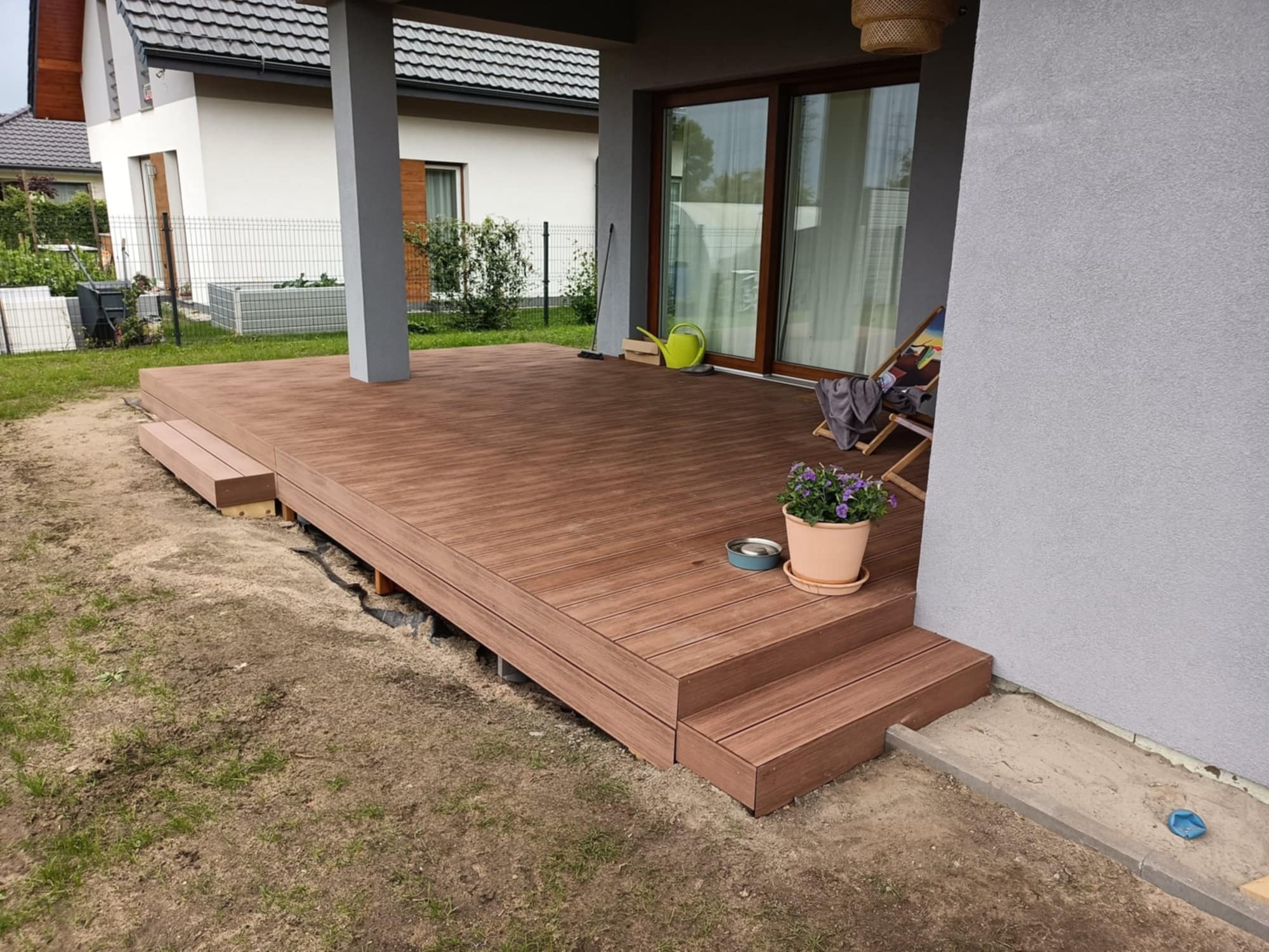 Nowoczesny taras z desek kompozytowych w kolorze brązu, z niskimi schodkami. Umeblowany, z widokiem na ogród i dom. Zastosowano oświetlenie zewnętrzne.