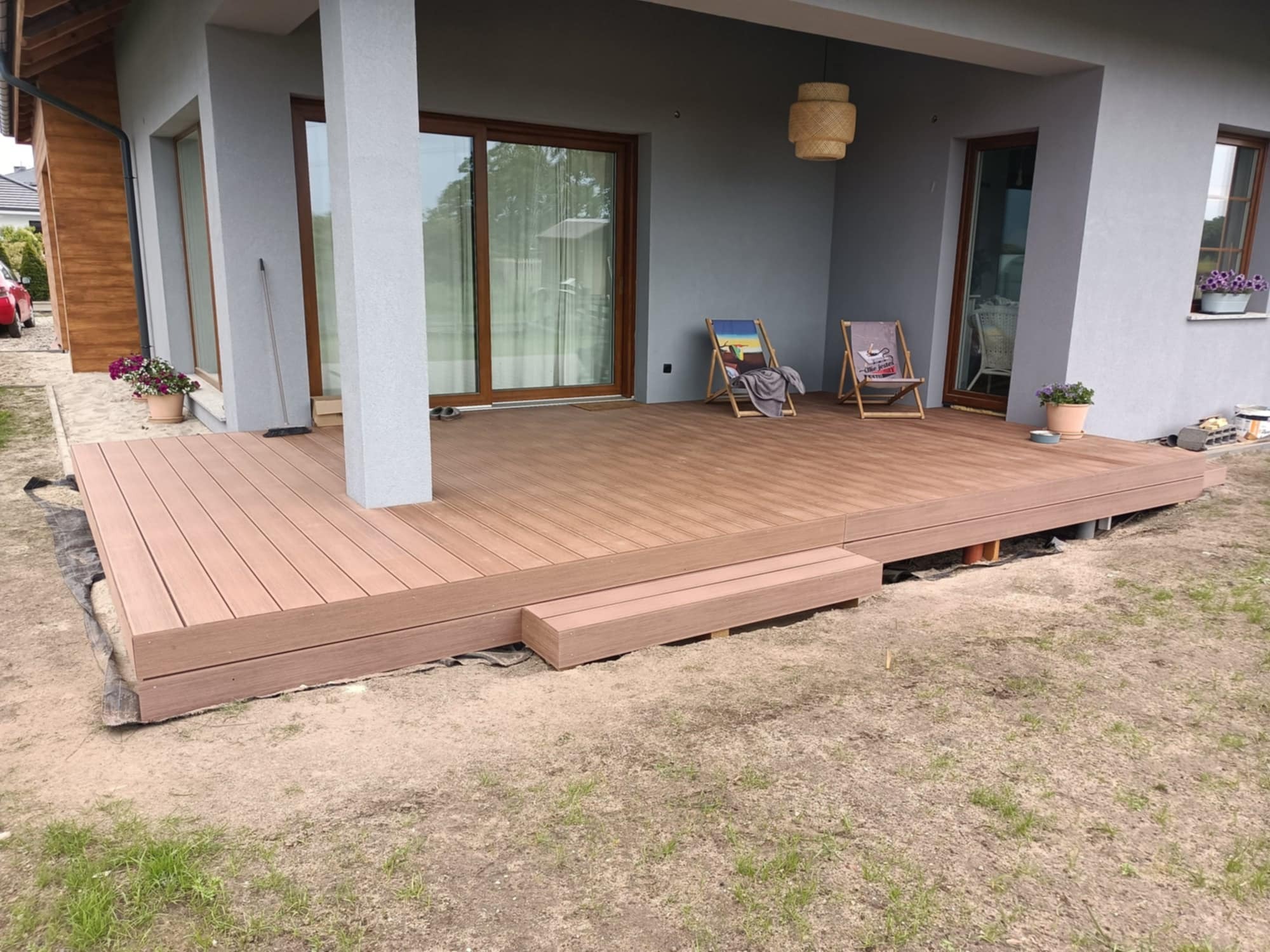 Nowoczesny taras kompozytowy z dwoma leżakami na tle szarego domu z dużymi oknami, oświetlony wiszącą lampą. Widoczne schodki wejściowe na taras.