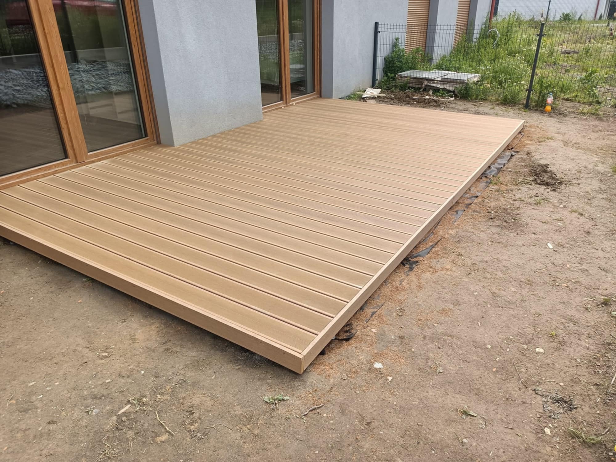 Beżowy taras kompozytowy przylegający do budynku z dużymi oknami, otoczony ziemią i zielenią. Widok z góry, jasne oświetlenie.