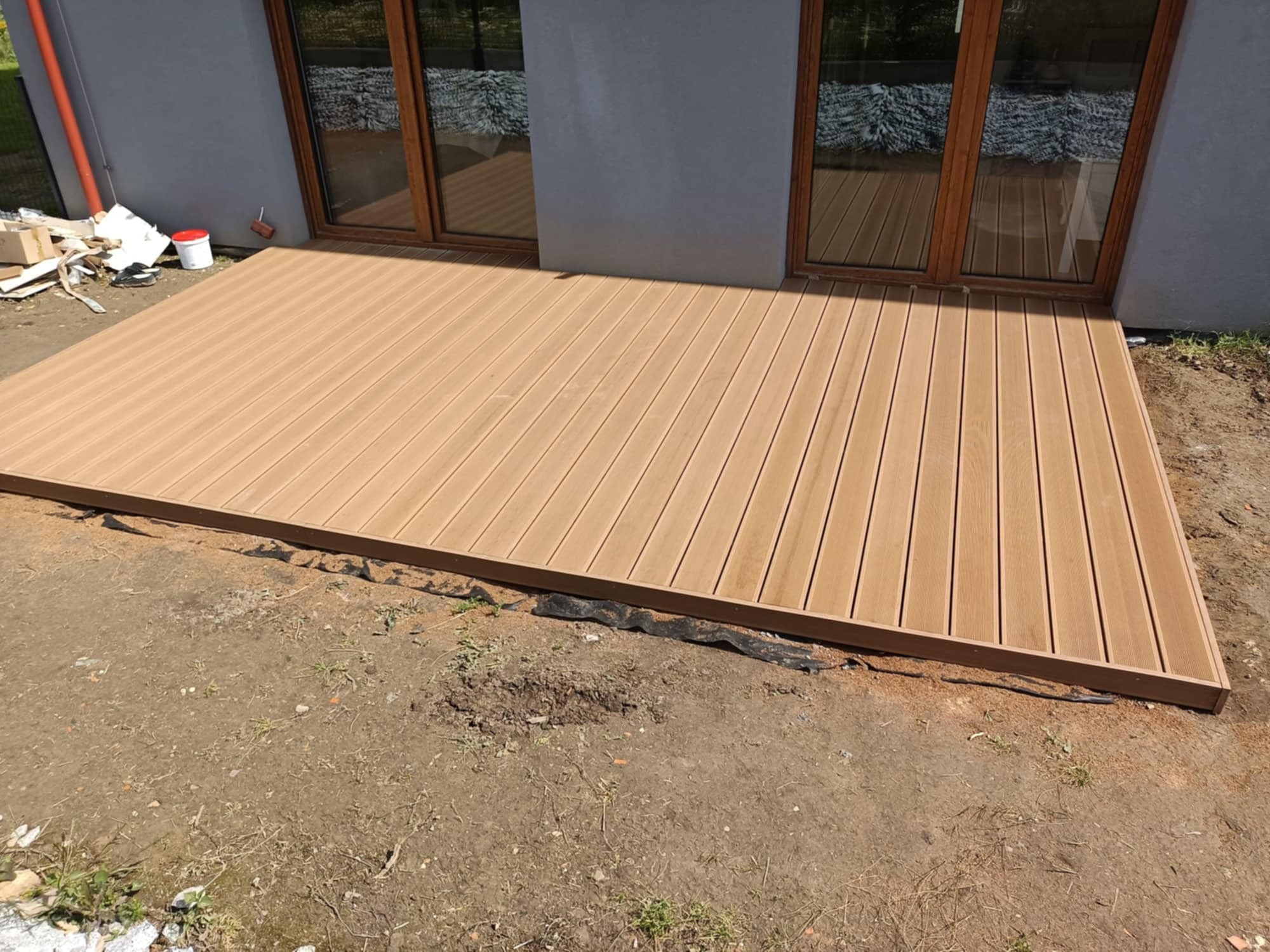 Jasny, kompozytowy taras przylegający do szarego budynku z drewnianymi oknami. Widoczne niedokończone obrzeża i ziemia wokół tarasu.