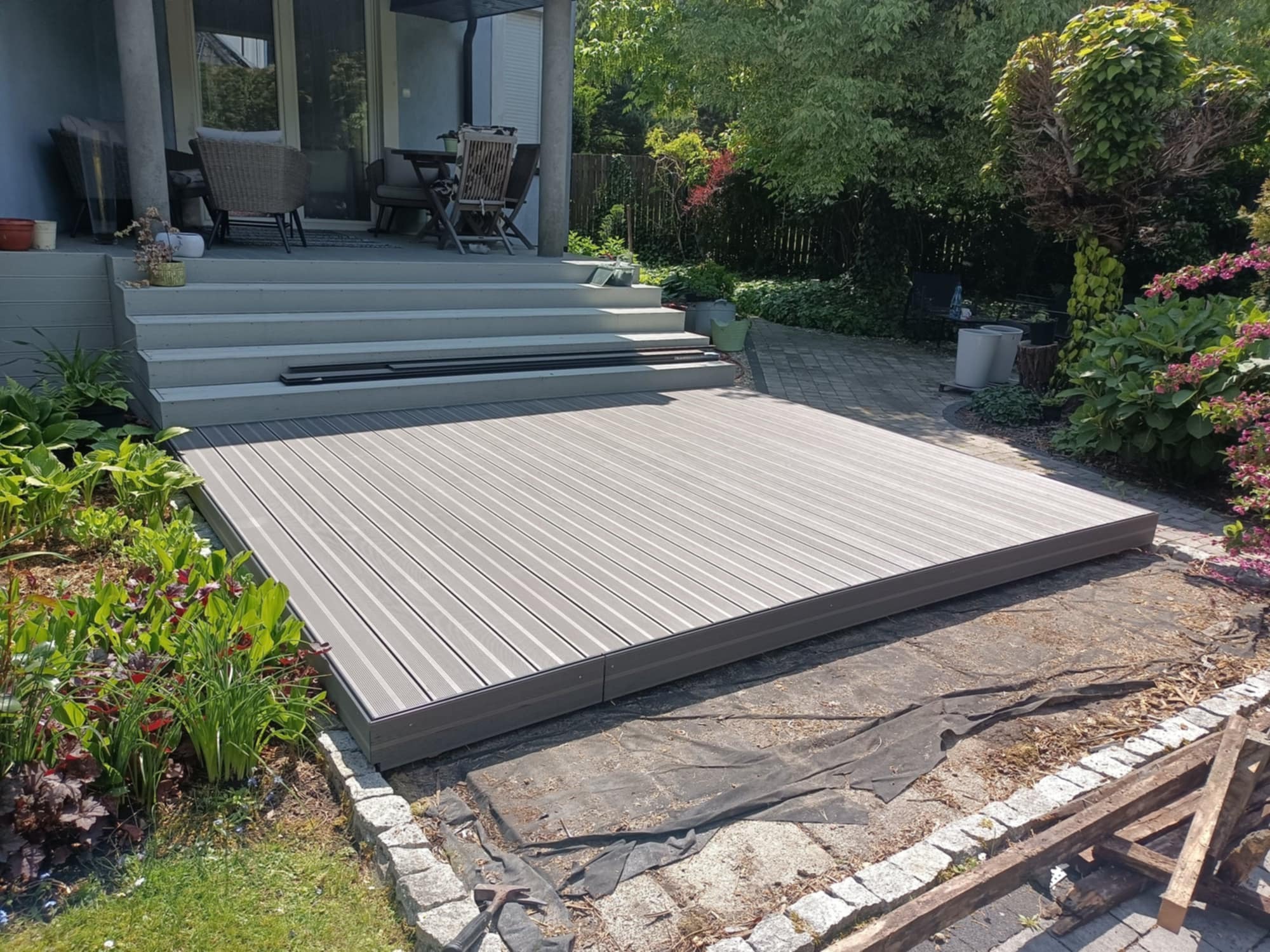 Montaż tarasu kompozytowego: Szare deski ułożone na przygotowanym podłożu, widoczne narzędzia i fragmenty starej nawierzchni. W tle schody i zieleń.