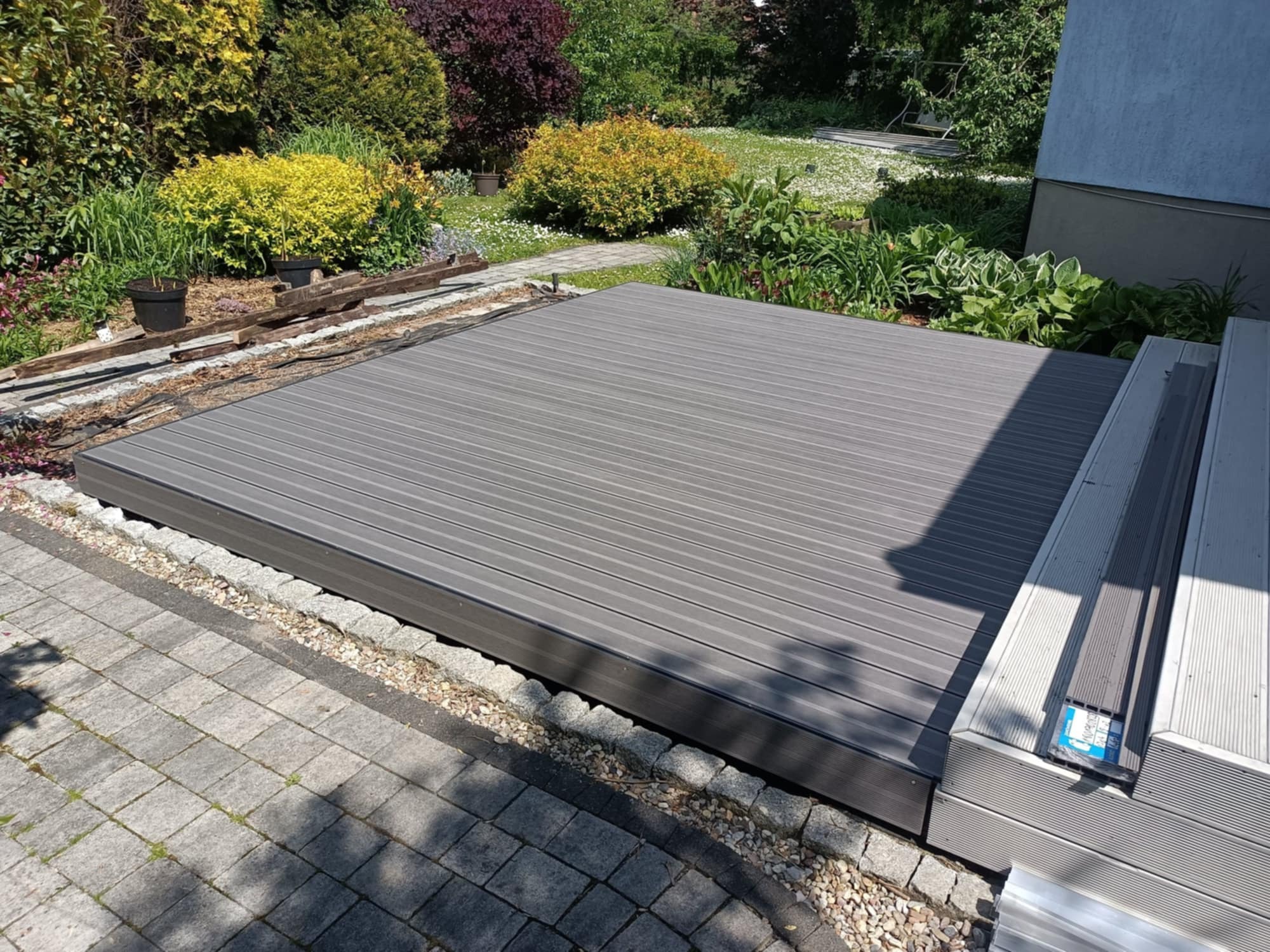 Nowoczesny taras kompozytowy w odcieniach szarości, otoczony zielenią ogrodu i kostką brukową. Widoczne detale wykończenia, naturalne światło słoneczne.