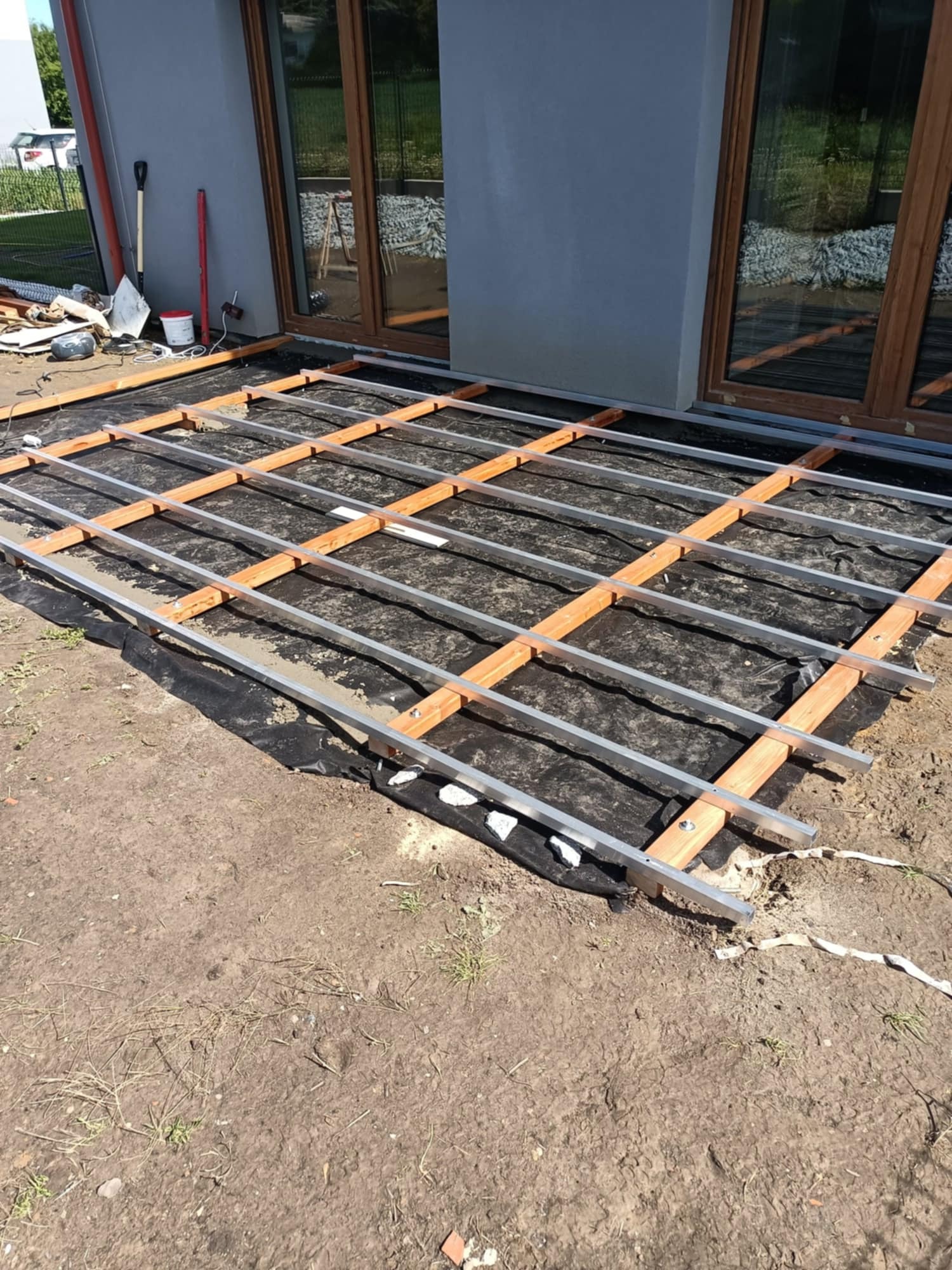 Konstrukcja tarasu: drewniane legary z aluminiowymi profilami na czarnej membranie, przygotowana pod deski. Widoczne narzędzia i elementy montażowe.