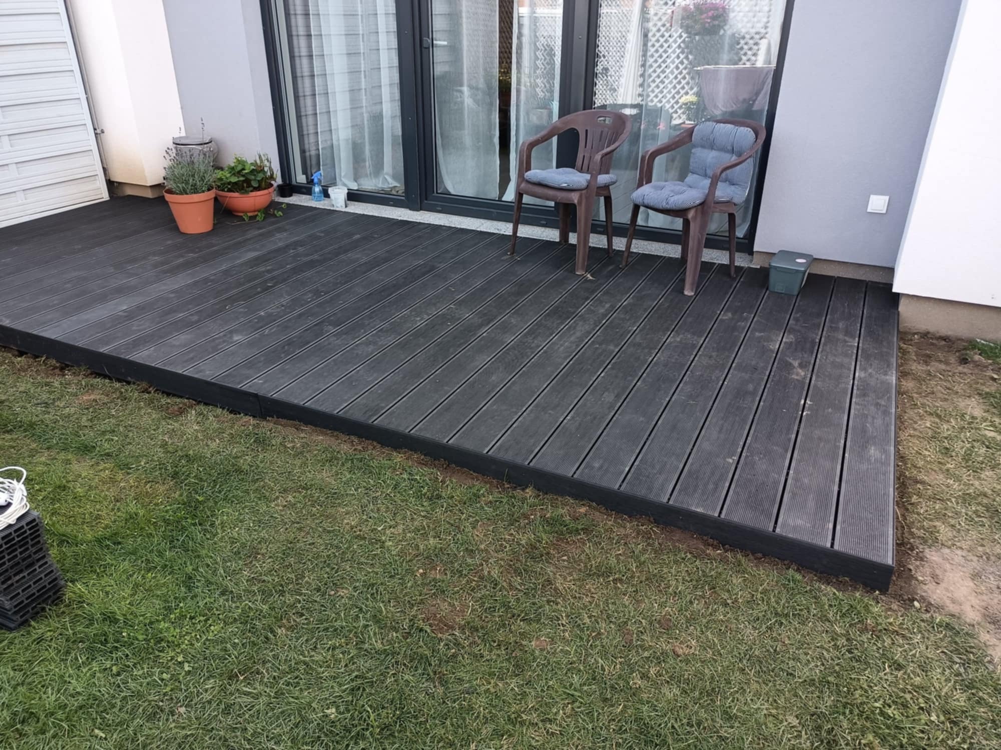Czarny taras kompozytowy z deskami ułożonymi wzdłuż, dwa krzesła z poduszkami, doniczki z roślinami przy oknie, trawnik. Widok z poziomu trawy.