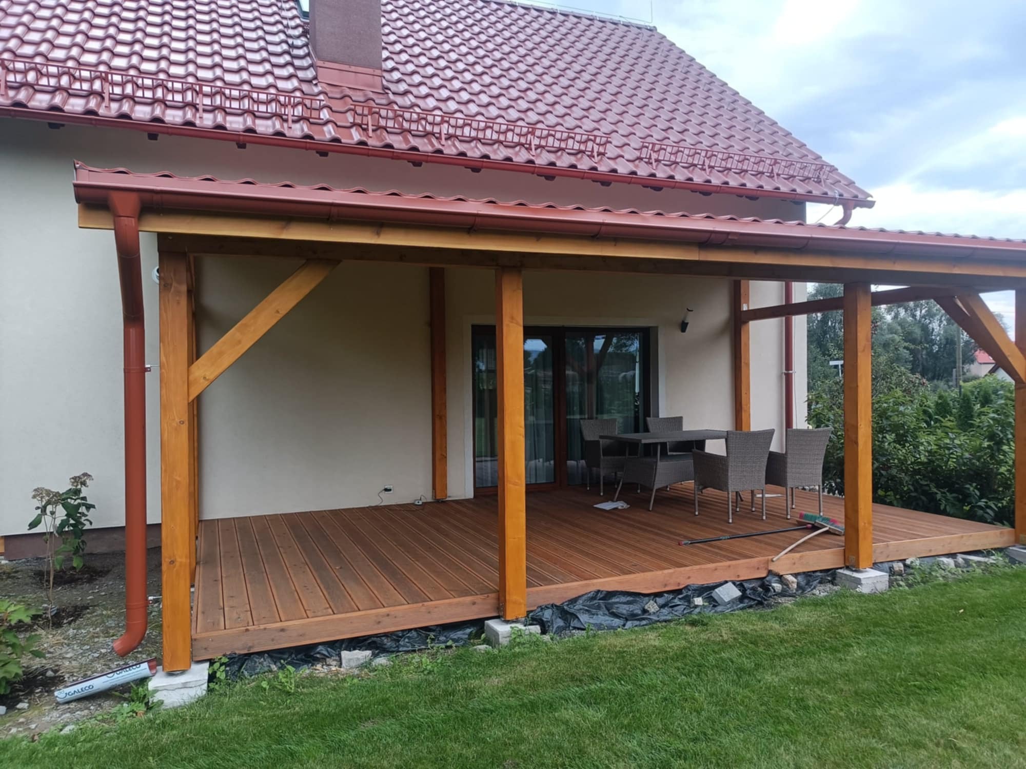 Zadaszony taras z drewna z kompletem mebli ogrodowych. Widoczne elementy konstrukcyjne i wykończeniowe, brązowa podłoga tarasu i fragment domu z czerwonym dachem.
