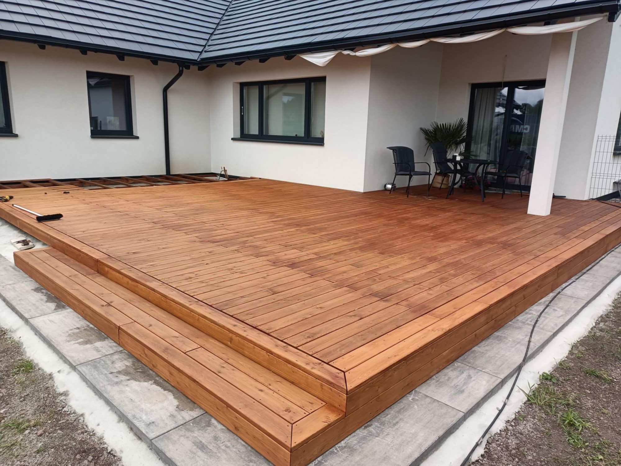 Nowoczesny taras drewniany z dwustopniowymi schodami, wykończony jasnym drewnem, przylegający do białego domu z ciemną stolarką okienną i zadaszeniem.