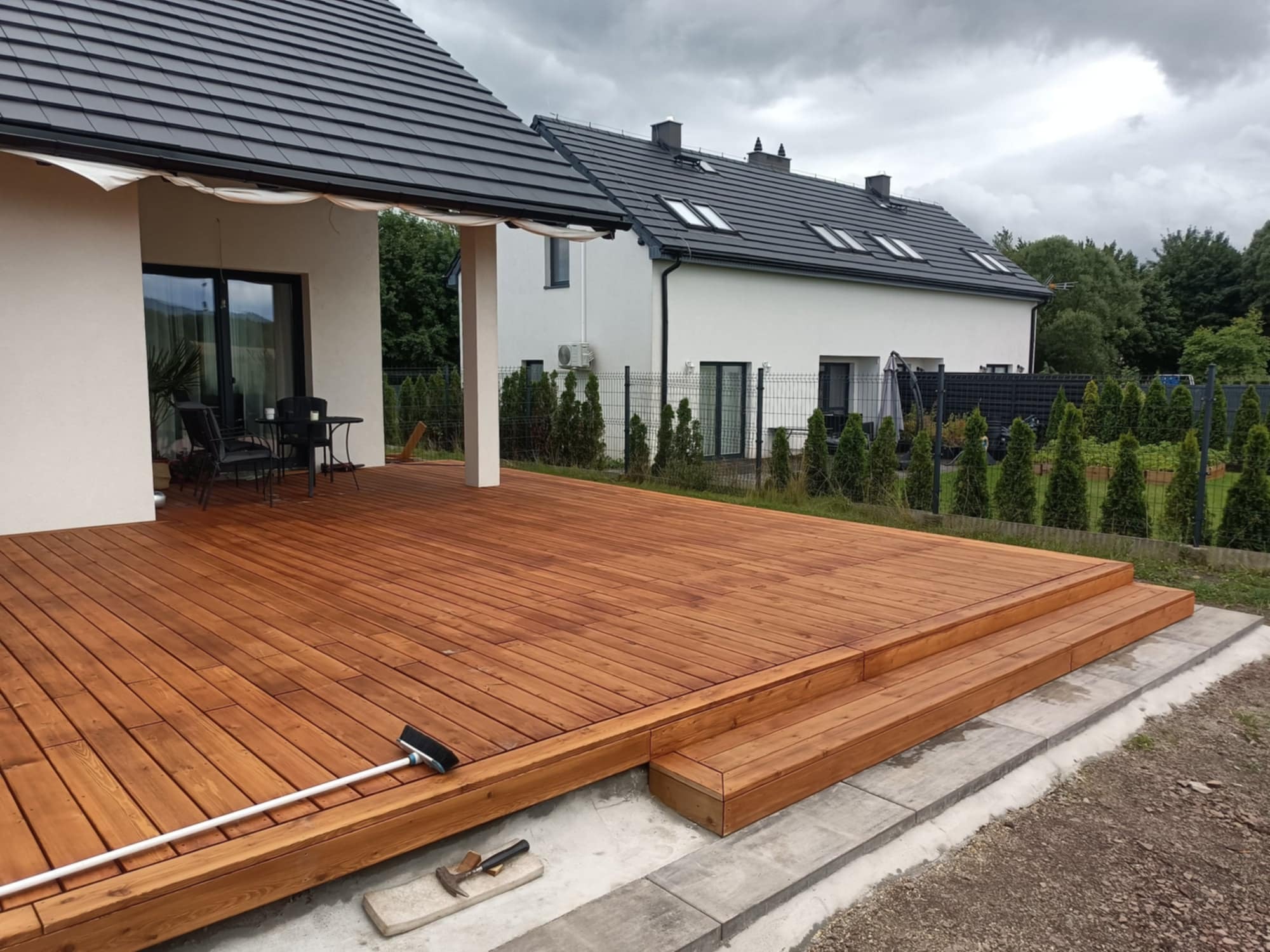 Świeżo wykończony taras z drewna egzotycznego, z widoczną szczotką do zamiatania i młotkiem na obrzeżu, przy nowoczesnym domu z szarą dachówką.