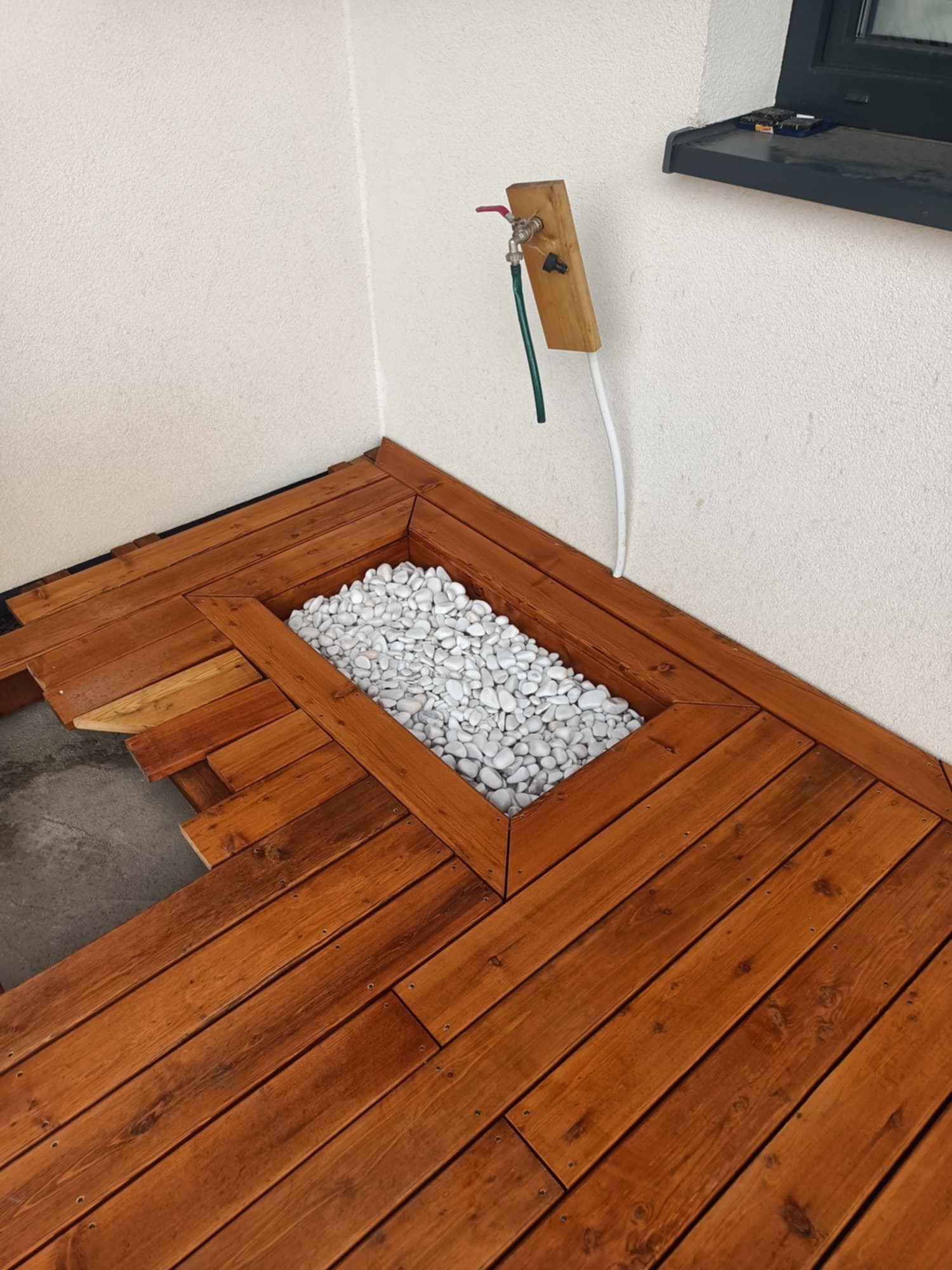 Brązowy taras drewniany w trakcie montażu, z wbudowaną donicą wypełnioną białymi kamieniami, widoczny kran z wężem na ścianie.