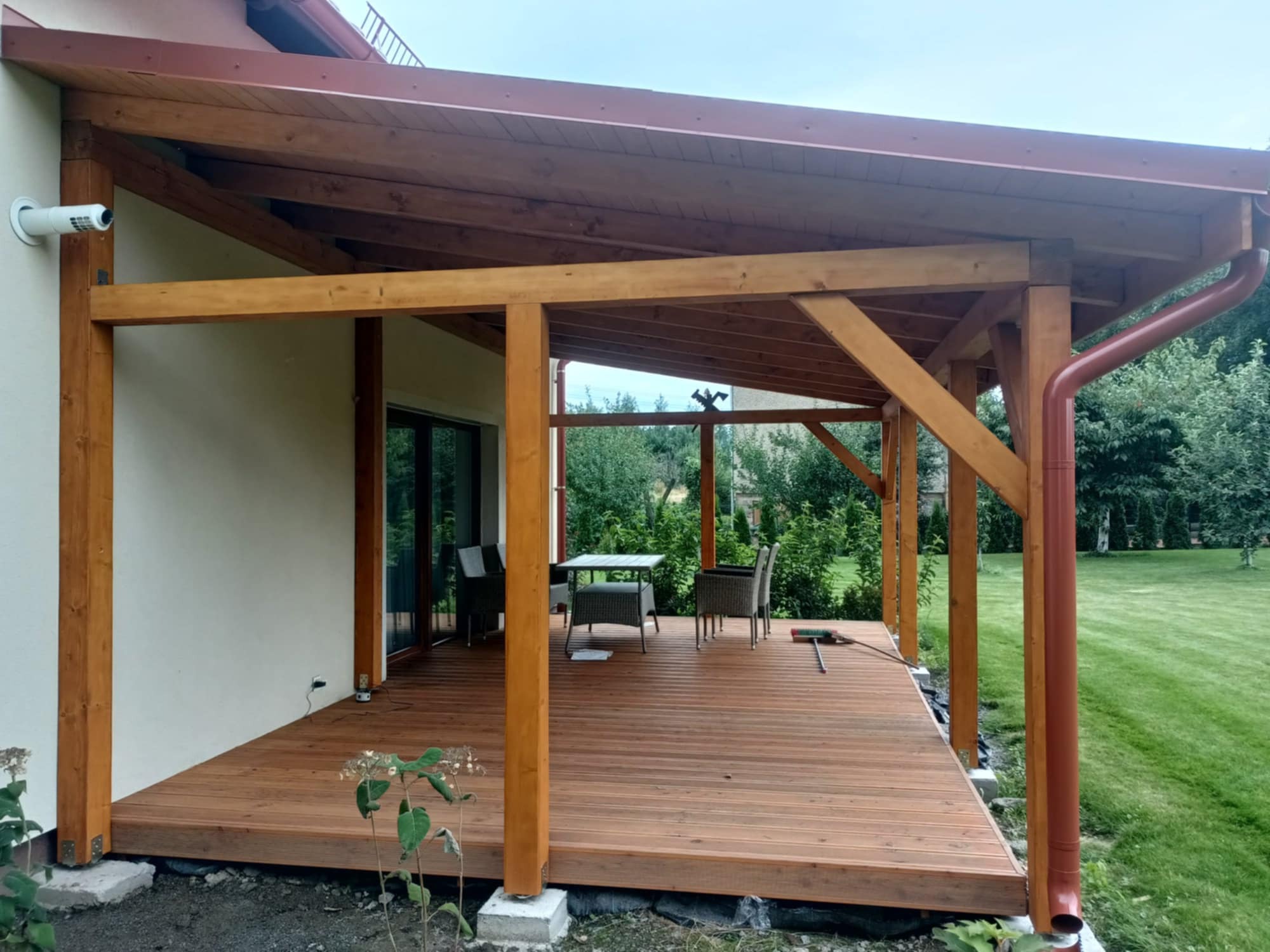 Zadaszony taras z drewnianą konstrukcją i meblami ogrodowymi. Widoczny system rynnowy w kolorze brązowym i kamera monitoringu na ścianie domu.