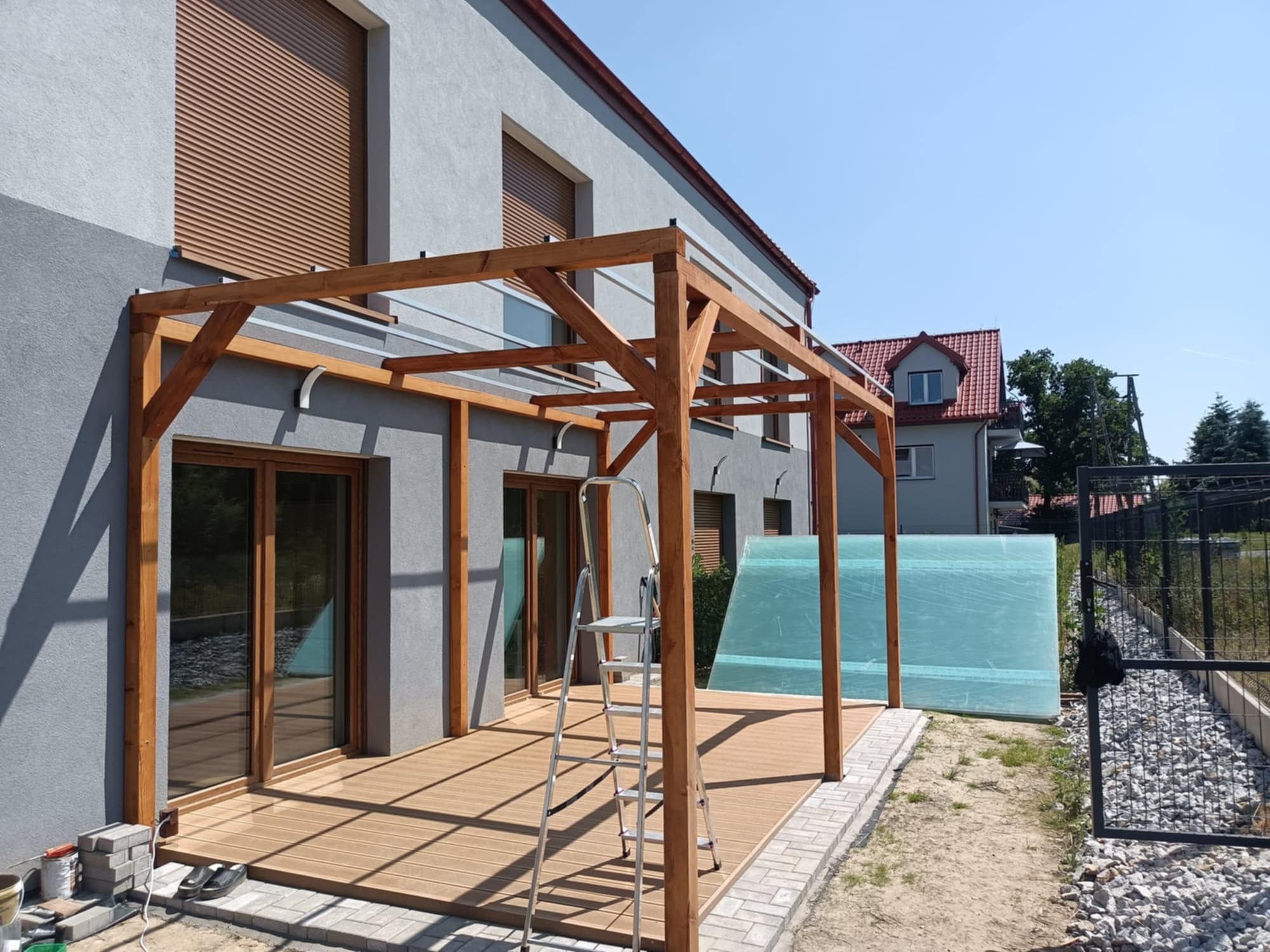 Drewniana pergola z zadaszeniem i tarasem kompozytowym przy szarym domu. Na tarasie stoi drabina. Widoczne materiały budowlane i fragment ogrodu.
