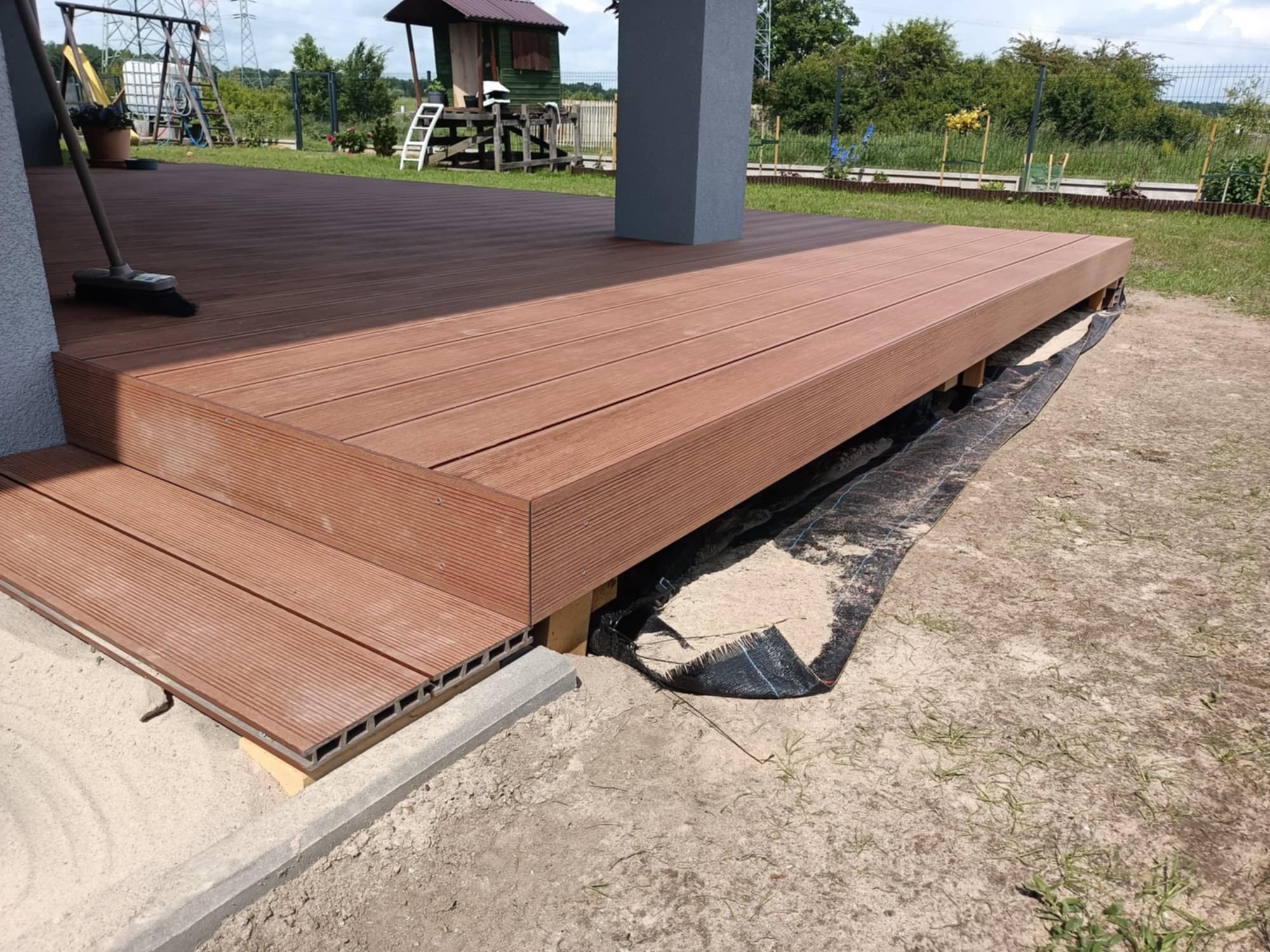 Gotowy taras kompozytowy w odcieniach brązu, z widocznymi deskami i schodami, na tle zieleni ogrodu. Ujęcie z niskiej perspektywy podkreśla strukturę materiału.