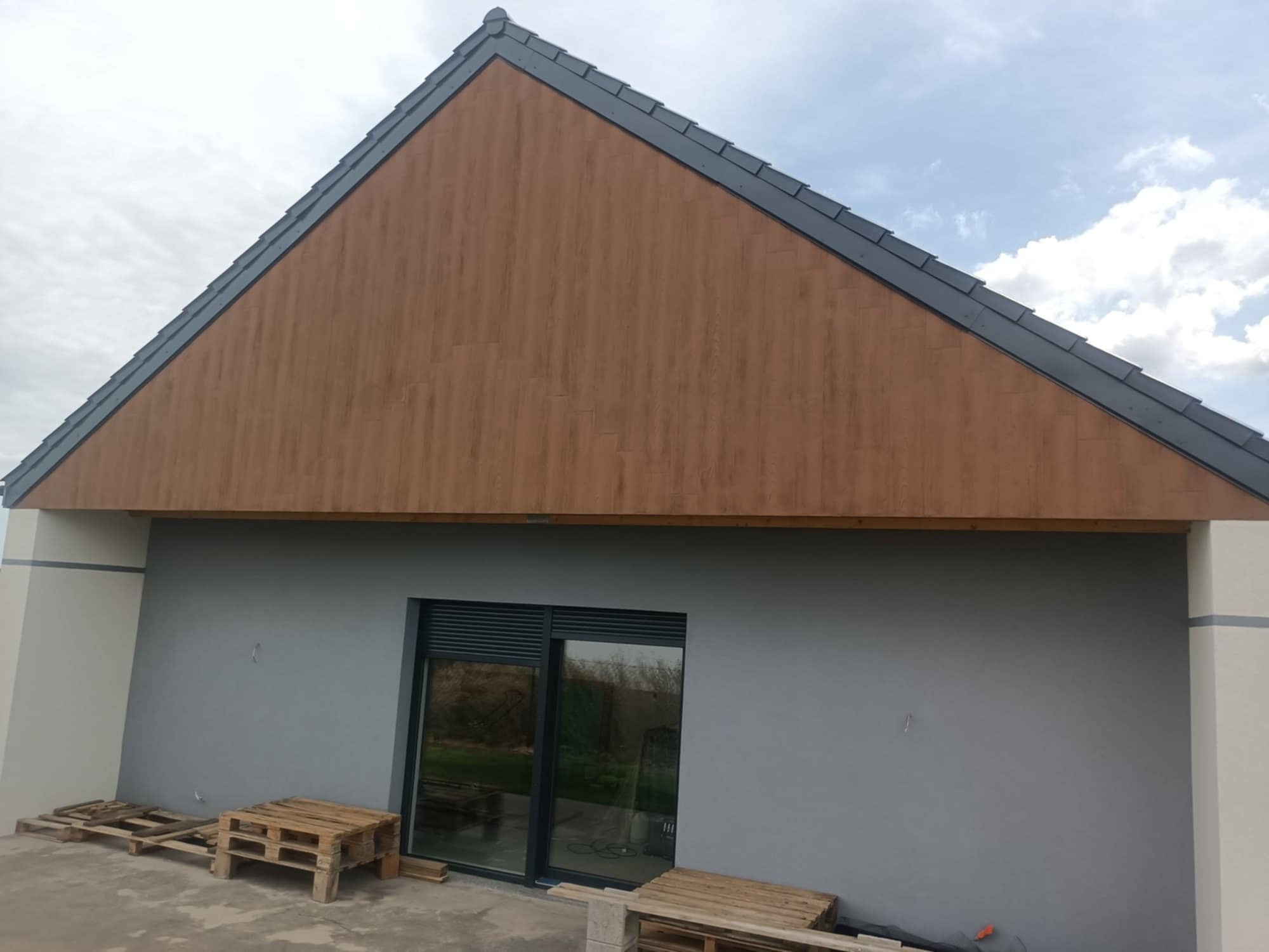 Wykończona elewacja domu z drewnopodobną okładziną szczytu i szarą ścianą. Widoczne okno, palety na ziemi. Ujęcie z dołu, pochmurne niebo w tle.