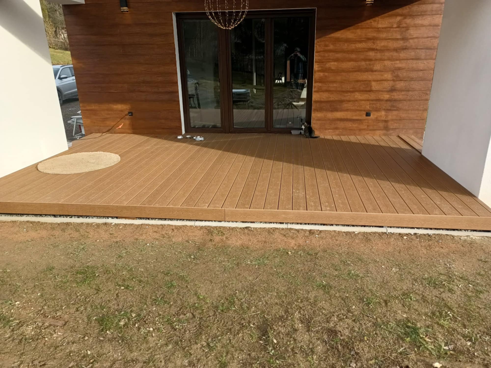Nowoczesny taras kompozytowy z brązowych desek, przylegający do budynku z drewnianą elewacją. Widoczny fragment trawnika i okrągły dywanik przy drzwiach.