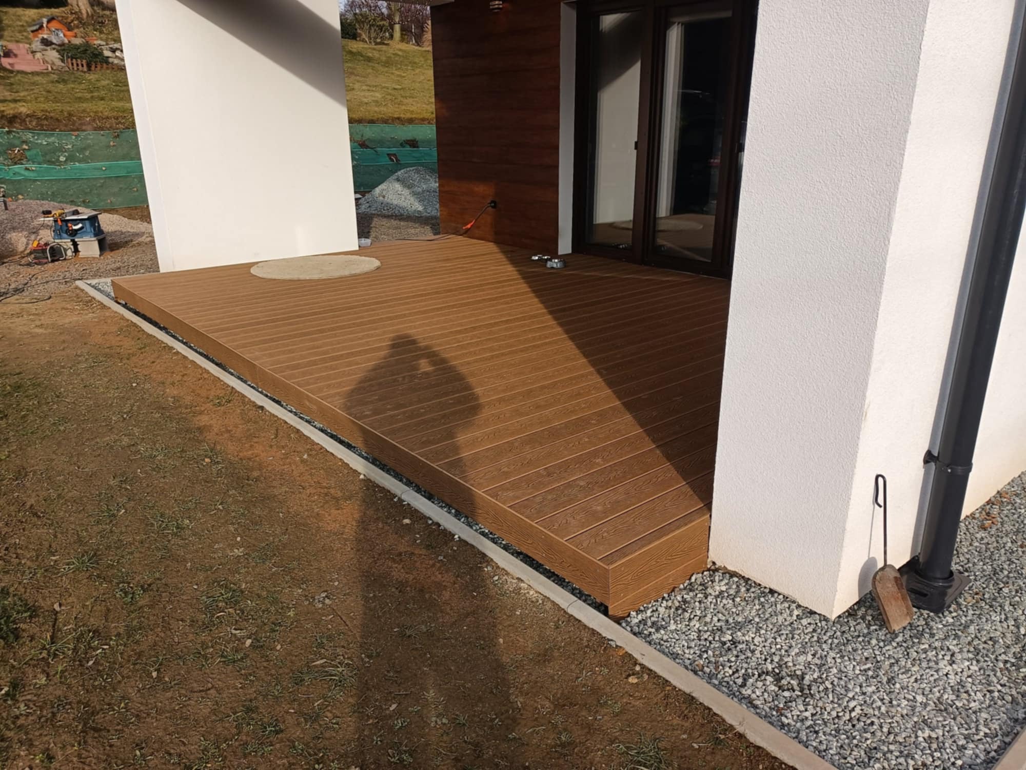 Nowoczesny taras z deski kompozytowej w kolorze brązowym, z widocznym cieniem osoby fotografującej, otoczony szarym żwirem i zielenią.