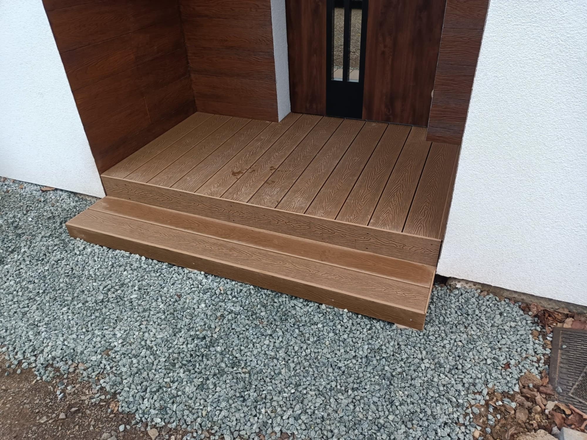 Wejście do domu z brązowego kompozytu. Schody i podest z desek imitujących drewno, otoczone białym murem i szarym żwirem. Ciemne drzwi z szybkami.