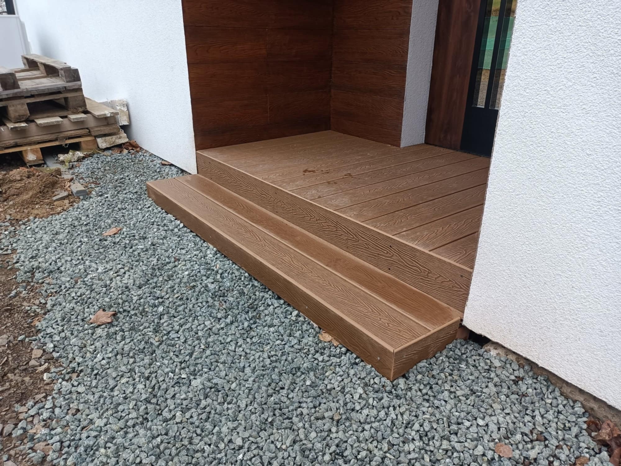 Nowoczesny taras z kompozytu w odcieniach brązu, z dwustopniowymi schodami, otoczony szarym żwirem i białą elewacją budynku. Widoczne deski imitujące drewno.