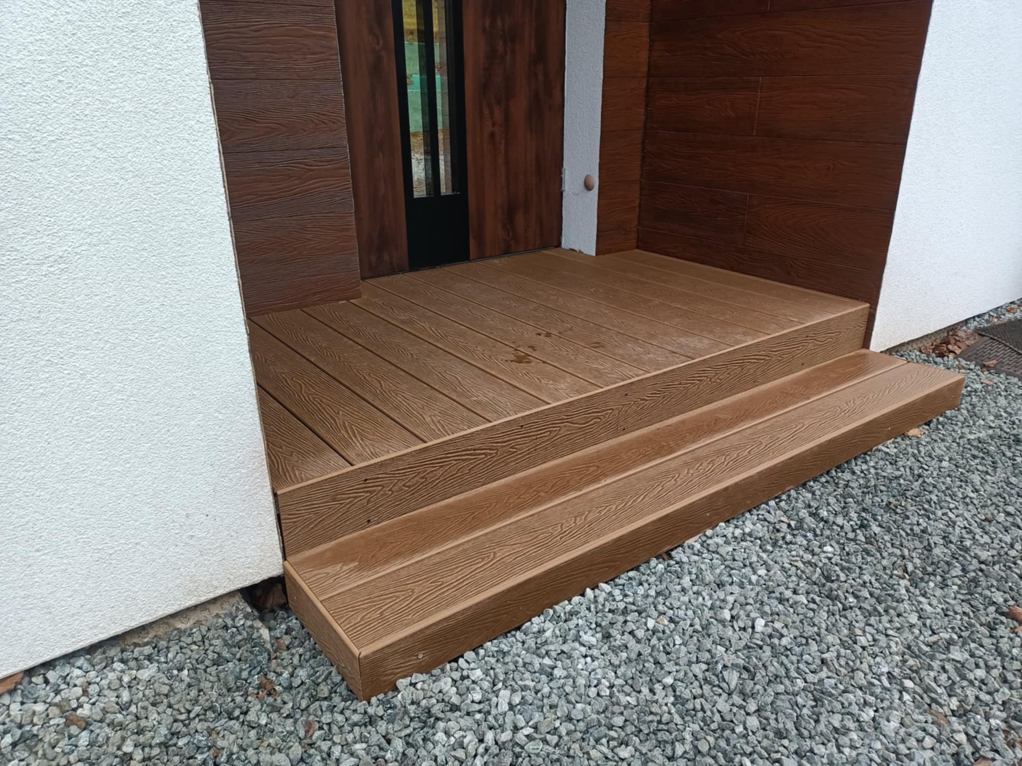 Brązowe schody z desek kompozytowych prowadzące do drzwi wejściowych, otoczone białym tynkiem i szarą żwirową nawierzchnią. Widoczne detale faktury desek.