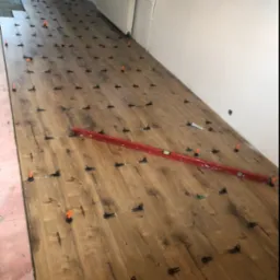 Układanie paneli podłogowych z użyciem systemu poziomowania, widoczna poziomica laserowa i kliny dystansowe.