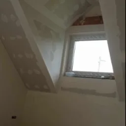 Poddasze w trakcie wykańczania: płyty karton-gips na skosach, szpachlowane łączenia, okno dachowe, kubek z kawą w dolnej części kadru.