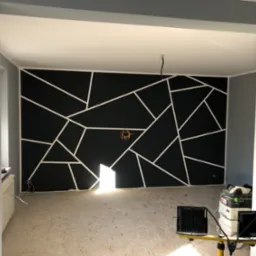 Pokój w trakcie remontu z pomalowaną na czarno ścianą, na której znajdują się białe, geometryczne wzory. Widoczne narzędzia i lampy budowlane, surowe podłoże i grzejnik.